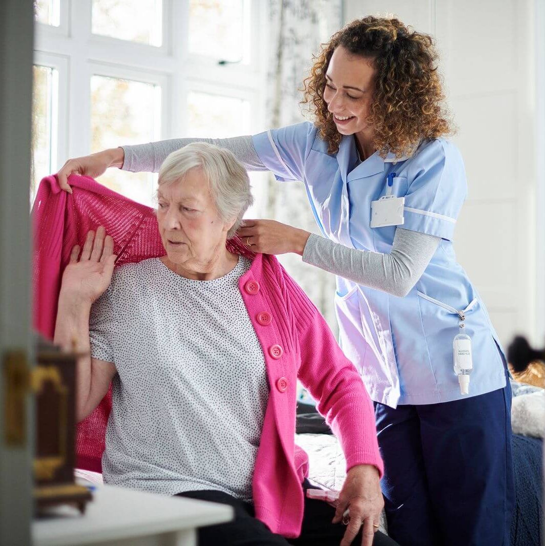 Nurse helping an elderly woman put on a cardigan during compassionate Home Care Support Services focused on daily personal care.